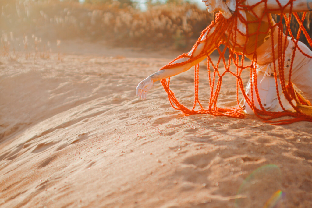 A dancer wearing a fire bird costume spreading her arm on her knees on a sand dune.